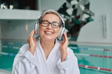 Gözlüklü ve bornozlu mutlu orta yaşlı kadın spa merkezinde kablosuz kulaklıkla müzik dinliyor.