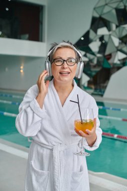 Mutlu orta yaşlı kadın kablosuz kulaklıkla müzik dinliyor ve spa merkezinde kokteyl tutuyor.