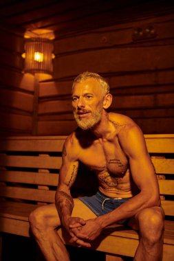 Rahatlamış ve gömleksiz orta yaşlı adam saunada oturuyor, sağIık inzivası konsepti.