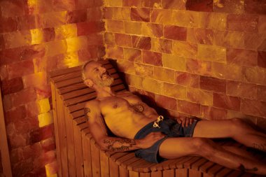 joyful and shirtless middle aged man with tattoos lying in sauna, wellness retreat concept clipart