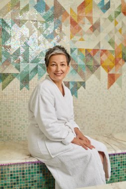 Beyaz cüppeli gülümseyen orta yaşlı kadın mermer saunada bankta oturuyor, spa sağlık konsepti.