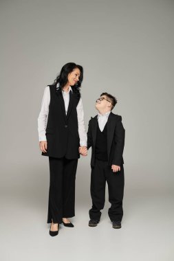 businesswoman and son with down syndrome holding hands, smiling at each other on grey, full length clipart