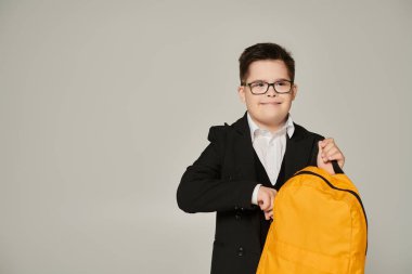 schoolboy with down syndrome holding yellow backpack and smiling on grey, inclusive schooling clipart