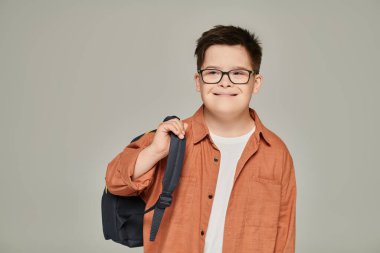 carefree schoolboy with mental disability holding backpack on grey, down syndrome concept clipart