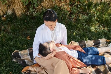 middle aged woman and son with down syndrome resting on blanket in park and smiling at each other clipart