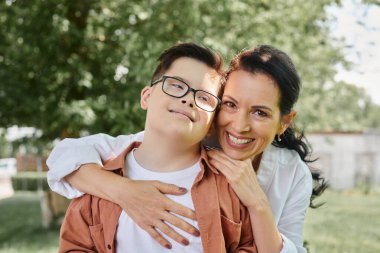 pleased middle aged woman embracing boy with down syndrome and smiling in park, special family clipart