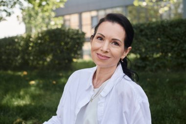 Parkta kameraya gülümseyen beyaz gömlekli orta yaşlı kadın portresi.