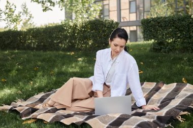 Yeşil parkta battaniyenin üzerinde oturan orta yaşlı kadın laptopta çalışıyor, serbest çalışıyor.