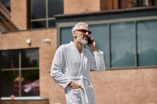 happy and tattooed middle aged man in sunglasses and robe talking on smartphone in resort, retreat