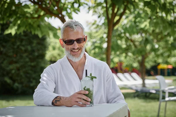 Sağlık sığınağı konsepti, güneş gözlüklü ve bornozlu mutlu orta yaşlı adam tatilde kokteylin tadını çıkarıyor.