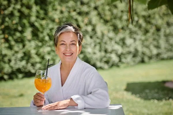 concept of wellness retreat, cheerful middle aged woman in robe enjoying cocktail on vacation