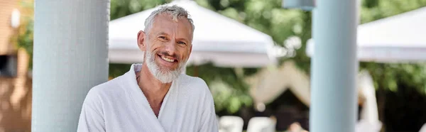 cheerful mature man in white robe looking at camera on resort, wellness retreat concept, banner