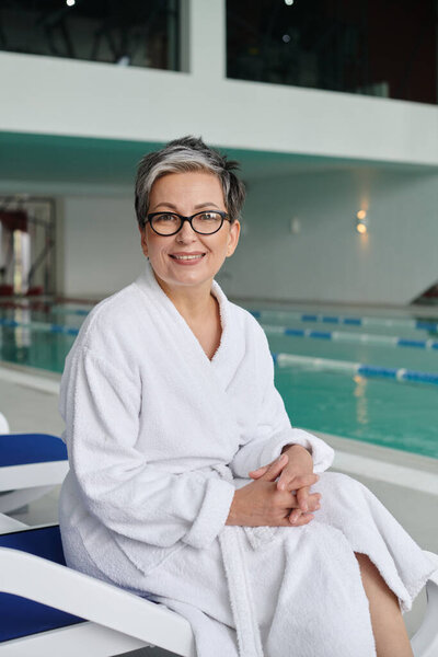 spa center concept, happy middle aged woman in glasses and robe sitting on lounger near pool