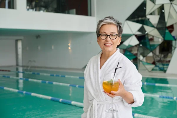 stock image spa center and retreat concept, happy mature woman in glasses and robe holding cocktail near pool