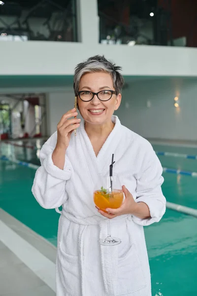 stock image wellness spa concept, happy mature woman in glasses holding cocktail and talking on smartphone