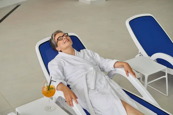 stock image retreat concept, pleased mature woman in white robe resting on lounger near cocktail in spa center