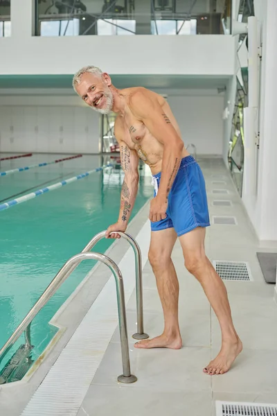 happy and shirtless mature man with tattoos and beard standing near pool ladder in spa center