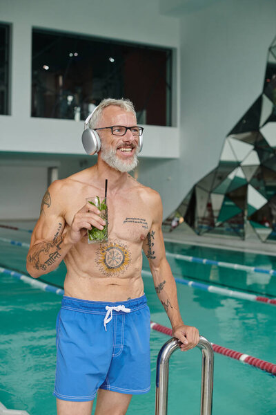 retreat, tattooed, happy mature man listening music in headphones and holding cocktail in spa