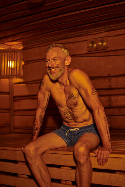 positive and shirtless middle aged man with tattoos sitting in sauna, wellness retreat concept