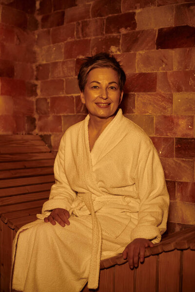 joyful middle aged woman in white robe sitting on bench in sauna, spa wellness concept, retreat