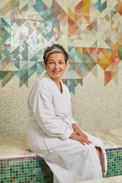 smiling middle aged woman in white robe sitting on bench in marble sauna, spa wellness concept