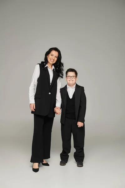 happy woman in formal wear holding hand of son with down syndrome in school uniform on grey