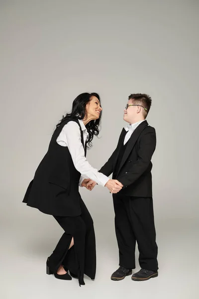 stock image joyful businesswoman holding hands of son with down syndrome in school uniform on grey, full length