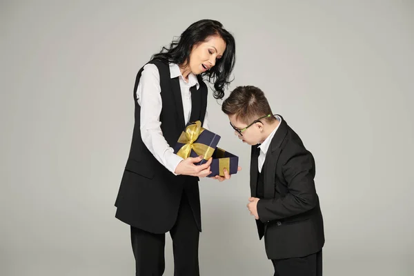 woman in formal wear presenting gift to surprised son with down syndrome in school uniform on grey