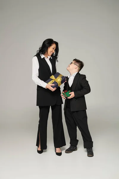 businesswoman and happy son with down syndrome in school uniform holding gift boxes on grey