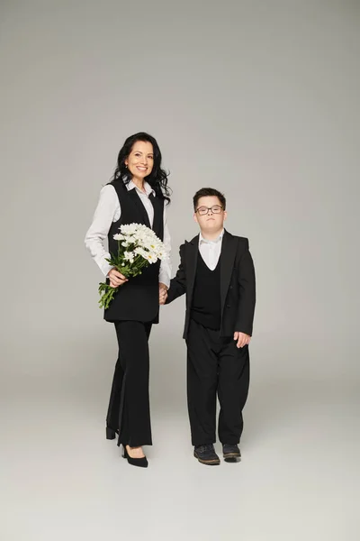 woman in formal wear holding flowers near son with down syndrome in school uniform on grey