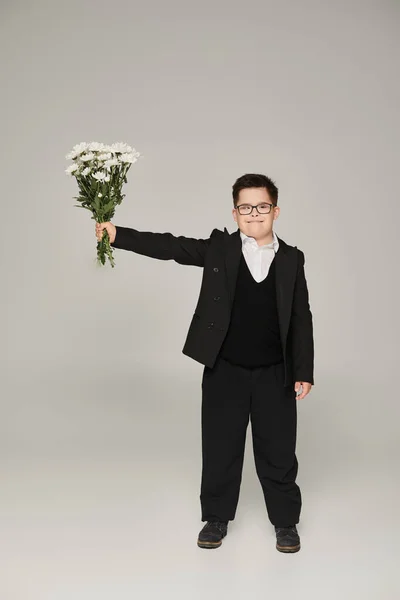 stock image schoolboy with down syndrome in eyeglasses and uniform holding bouquet in outstretched hand on grey