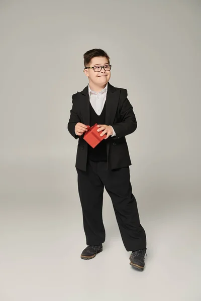 stock image cheerful boy with down syndrome in school uniform and eyeglasses standing with red gift box on grey