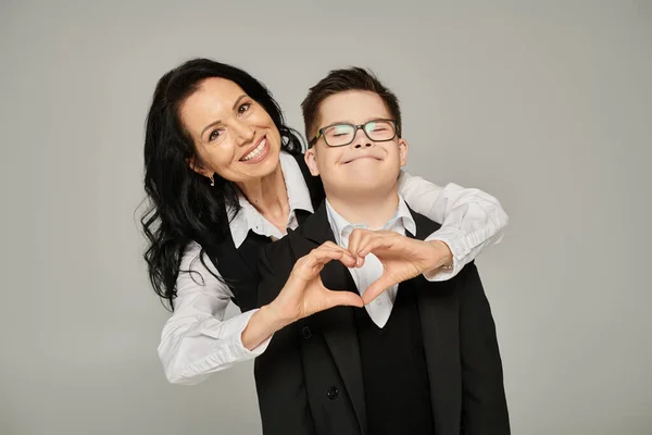 happy woman in formal wear showing love sign near son with down syndrome in school uniform on grey