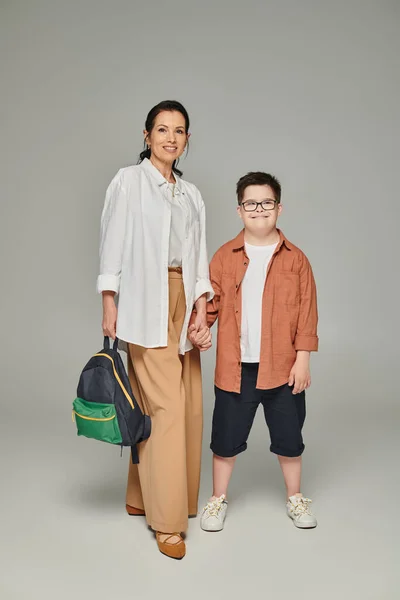 middle aged woman with school backpack and son with down syndrome holding hands on grey, full length