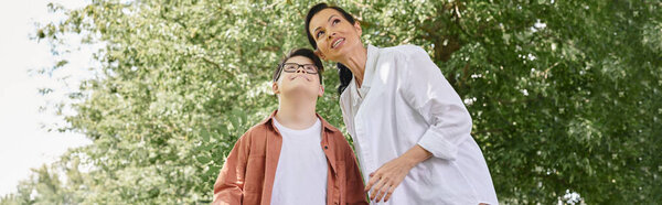 preteen boy with down syndrome near smiling mother looking away while walking in park, banner