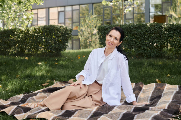cheerful middle aged woman in trendy casual attire sitting on blanket in park and smiling at camera