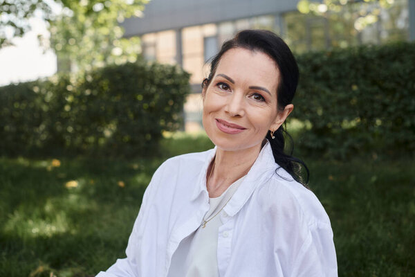 portrait of positive and stylish middle aged woman in white shirt smiling at camera in park