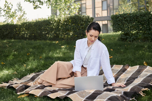 middle aged woman in casual attire sitting on blanket in green park and working on laptop, freelance