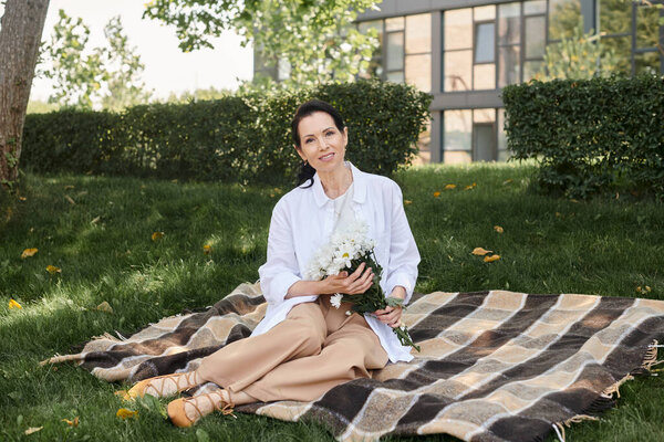cheerful and stylish middle aged woman with flowers sitting on blanket in park and looking at camera