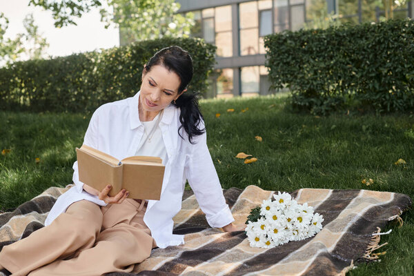 smiling middle aged woman sitting on blanket near flowers and reading book in green park
