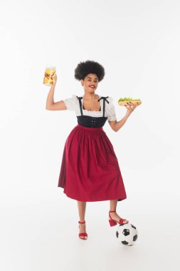 Mutlu Afro-Amerikan Bavyera garsonu Dirndl 'de bira kupası, sosisli sandviç ve futbol topuyla