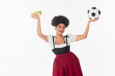 excited african american bavarian waitress with soccer ball and hot dog in raised hands on white clipart