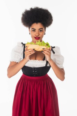 african american oktoberfest waitress in bavarian costume holding hot dog and smiling on white clipart