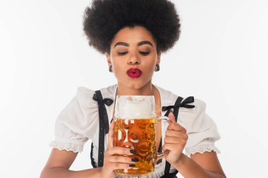 Oktoberfest kostümlü Afro-Amerikan Bavyera garsonu beyaz bardakta bira köpüğü üflüyor.