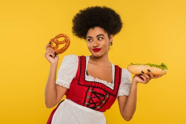 happy african american oktoberfest waitress in traditional dress with hot dog and pretzel on yellow clipart