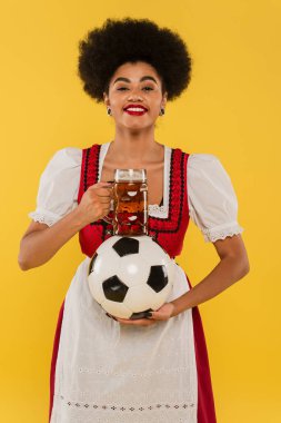 Bira bardağı ve futbol topuyla sarıya gülümseyen mutlu Afro-Amerikan Bavyera garsonu