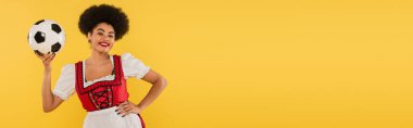 Mutlu Afro-Amerikan Bavyera garsonu futbol topuyla poz veriyor ve elini sarıya koyuyor.