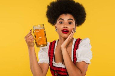 Şaşırmış Afro-Amerikan Bavyera garsonu bira bardağı ve açık ağzıyla sarıya dokunuyor.