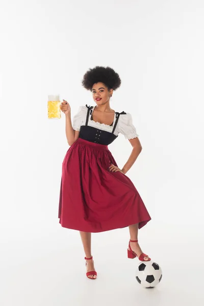 stock image happy african american bavarian waitress posing with mug of beer with foam near soccer ball on white