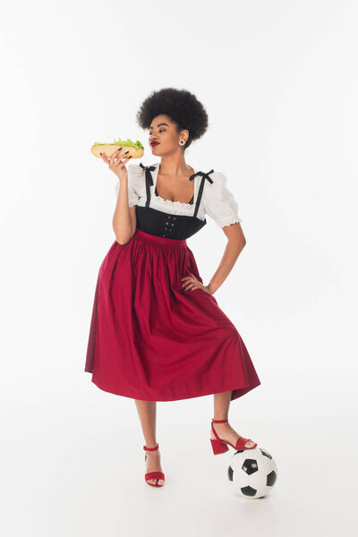 african american bavarian waitress posing with tasty hot dog and soccer ball on white, oktoberfest